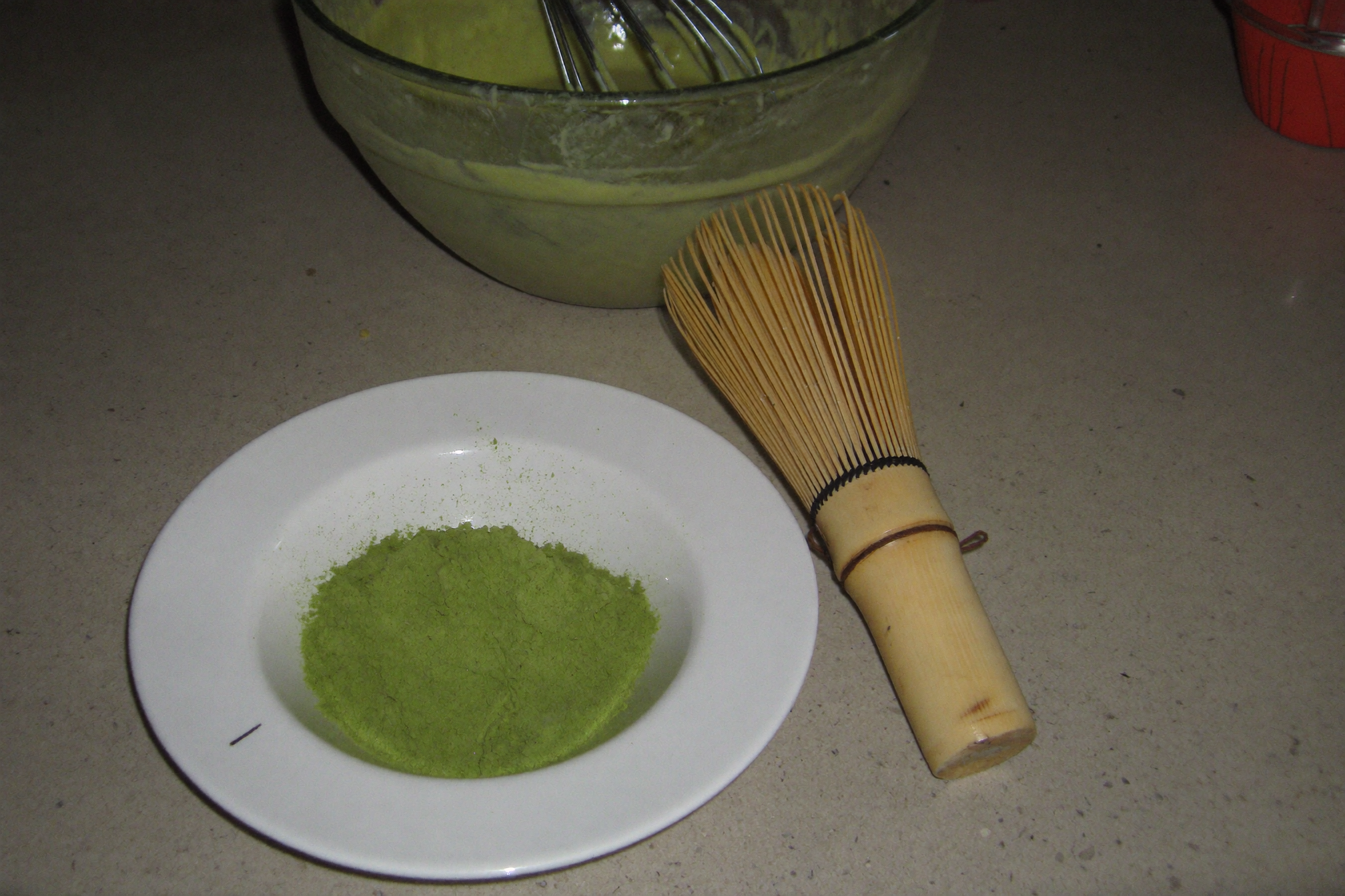 Dish of ceremonial matcha powder beside a bamboo whisk with culinary matcha batter behind