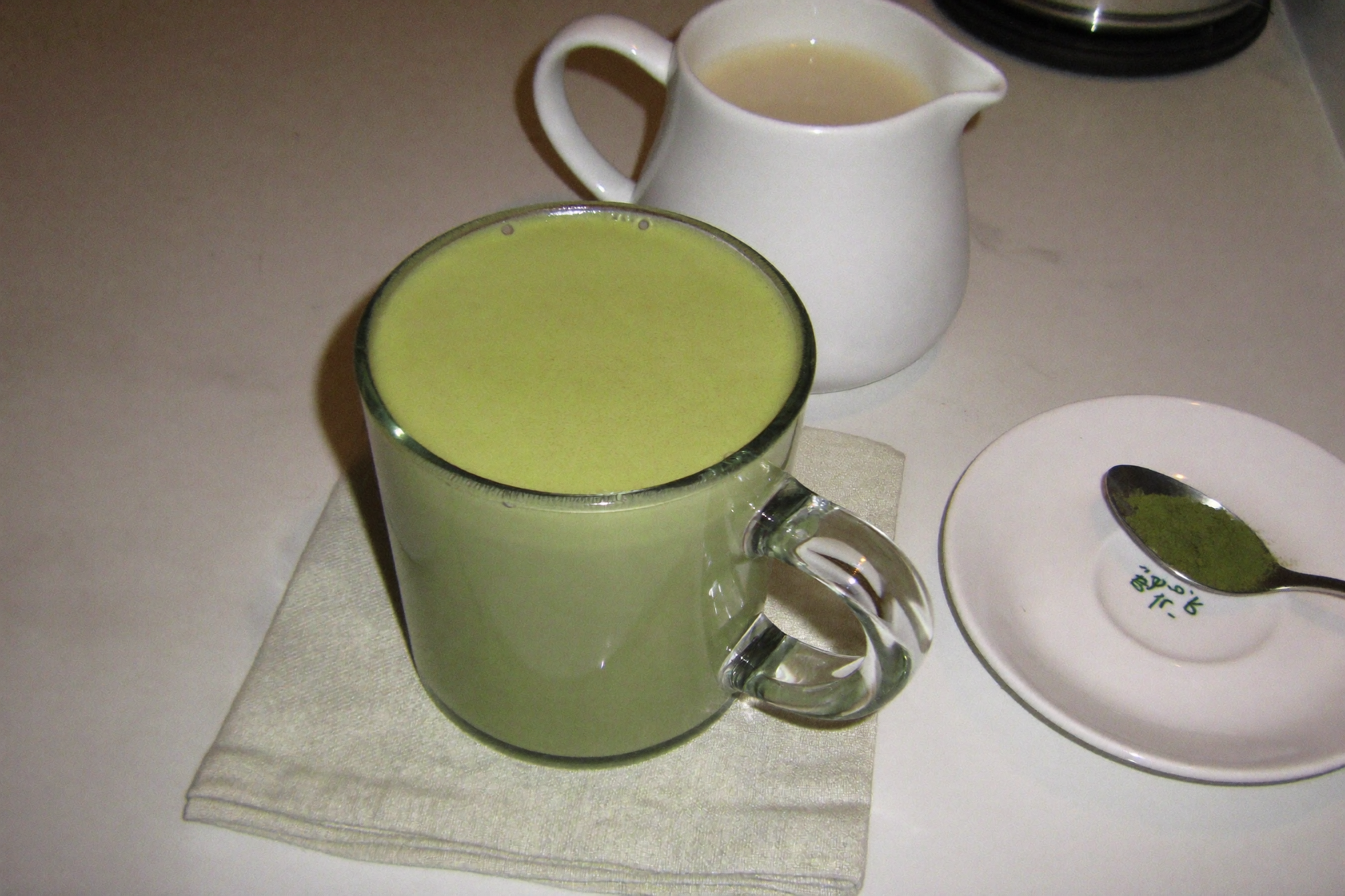 Homemade hot matcha latte in a glass mug with a milk jug and spoon of matcha powder